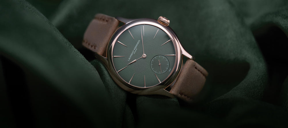 Lifestyle front view of the Laurent Ferrier Classic 'Evergreen' with red gold case. The watch with a brown bracelet is laying on its side on a crumpled forest green matte satin fabric.