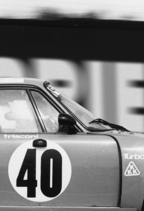 Side black and white in motion profile of racing number 40 car in Le Mans, France, 24 hours race driven by friends Laurent Ferrier and François Servanin. The car features sponsors stickers and is against a motion blurred background of ads.