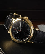 laurent ferrier ecole micro rotor yellow gold case, black opaline dial, red accents and white assegai hands, black bracelet, watch laying on it's side on a black leather notebook