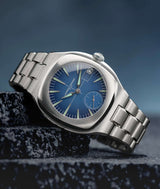 commercial ad visual of laurent ferrier fine watchmaking geneva's sport auto watch with titanium grade 5 included bracelet, blue gradient dial and mint green superluminova white gold hands and indexes, the timepiece is resting on its side on a pile of black car racetrack tar and smokey blue background. photographer cyril biselx