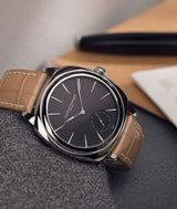 Laurent Ferrier square cushion shaped watch in stainless steel with beige light tan alligator bracelet and black opaline dial resting on grey fabric with pen and paper in background